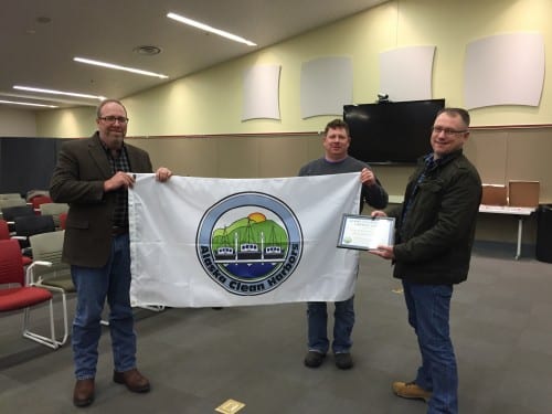 From L to R: Chuck Hackett, Brett Farrell, and Stan Eliason hold the flag, designating Sitka as an "Alaskan Clean Harbor." (Emily Kwong/KCAW photo)