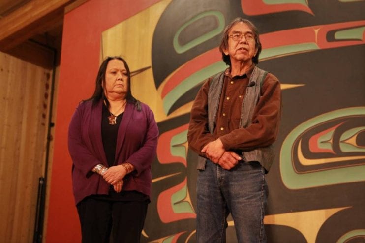 Tribal Council member Bob Sam stood on stage at the Sheet'ka Kwan Naa Kahidi by Louise Brady's side as he told the dining crowd the oral story of the origins of herring harvesting. (KCAW photo/Enrique Pérez de la Rosa)